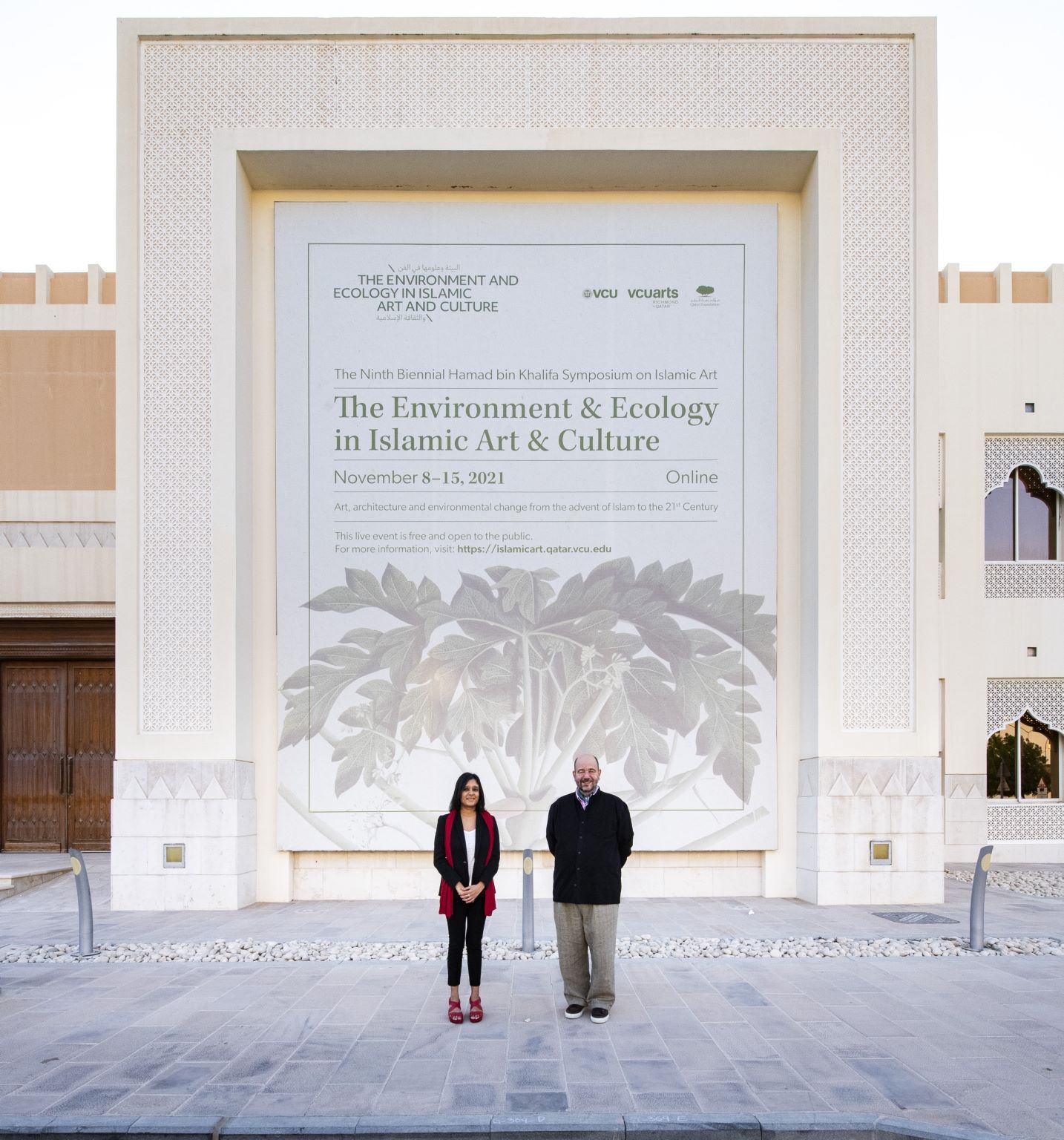 Radha Dalal and Jochen Sokoly standing in front of the branding for The Environment and Ecology in Islamic Art and Culture which was held at V C U arts Qatar in 2021