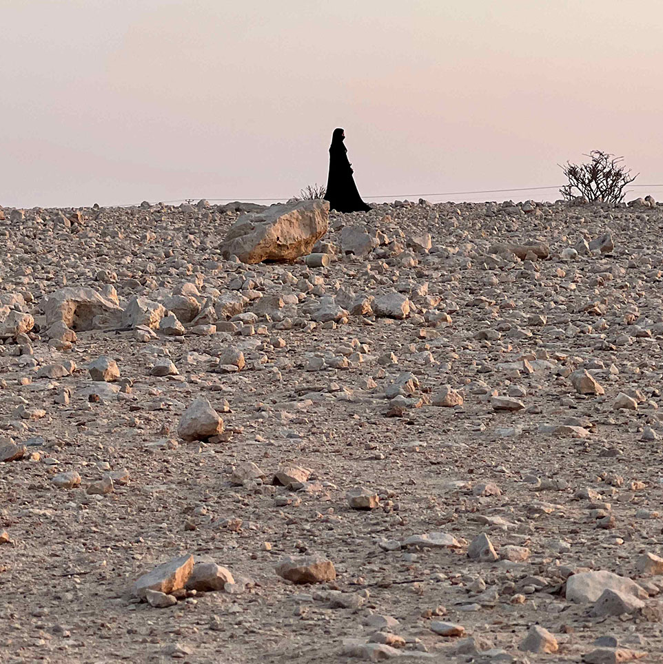 Photograph of a woman wearing an abaya walking through a rocky desert 