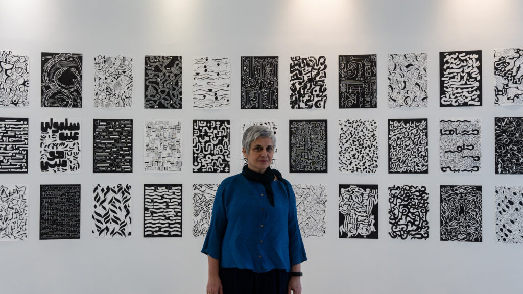 Huda Standing In Front Of More Than Thirty Monochromatic Posters On A Wall