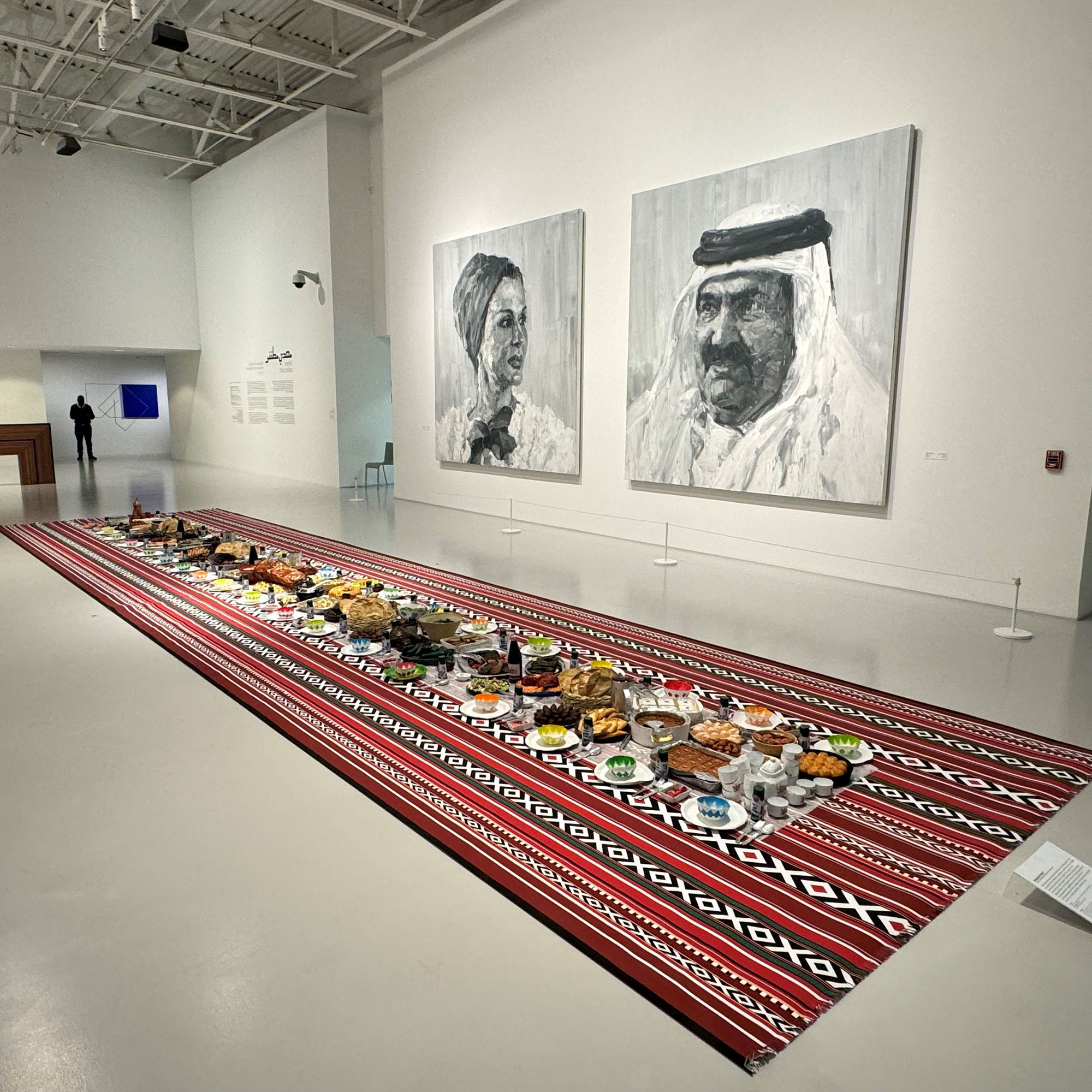 The Ramadan Feast work on display at Mathaf . In the background there are two paintings . One is of His Highness The Father Amir Sheikh Hamad bin Khalifa Al Thani . The other is of Her Highness Sheikha Moza bint Nasser