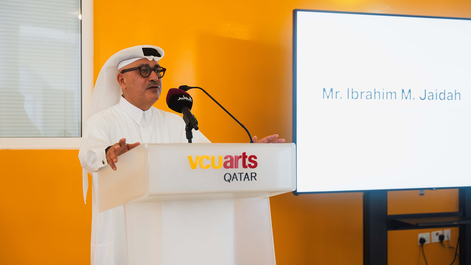 Ibrahim Mohamed Jaidah addressing the audience . He is standing at the podium with the words : V C U arts Qatar on the front . the screen behind him says Ibrahim Mohamed Jaidah