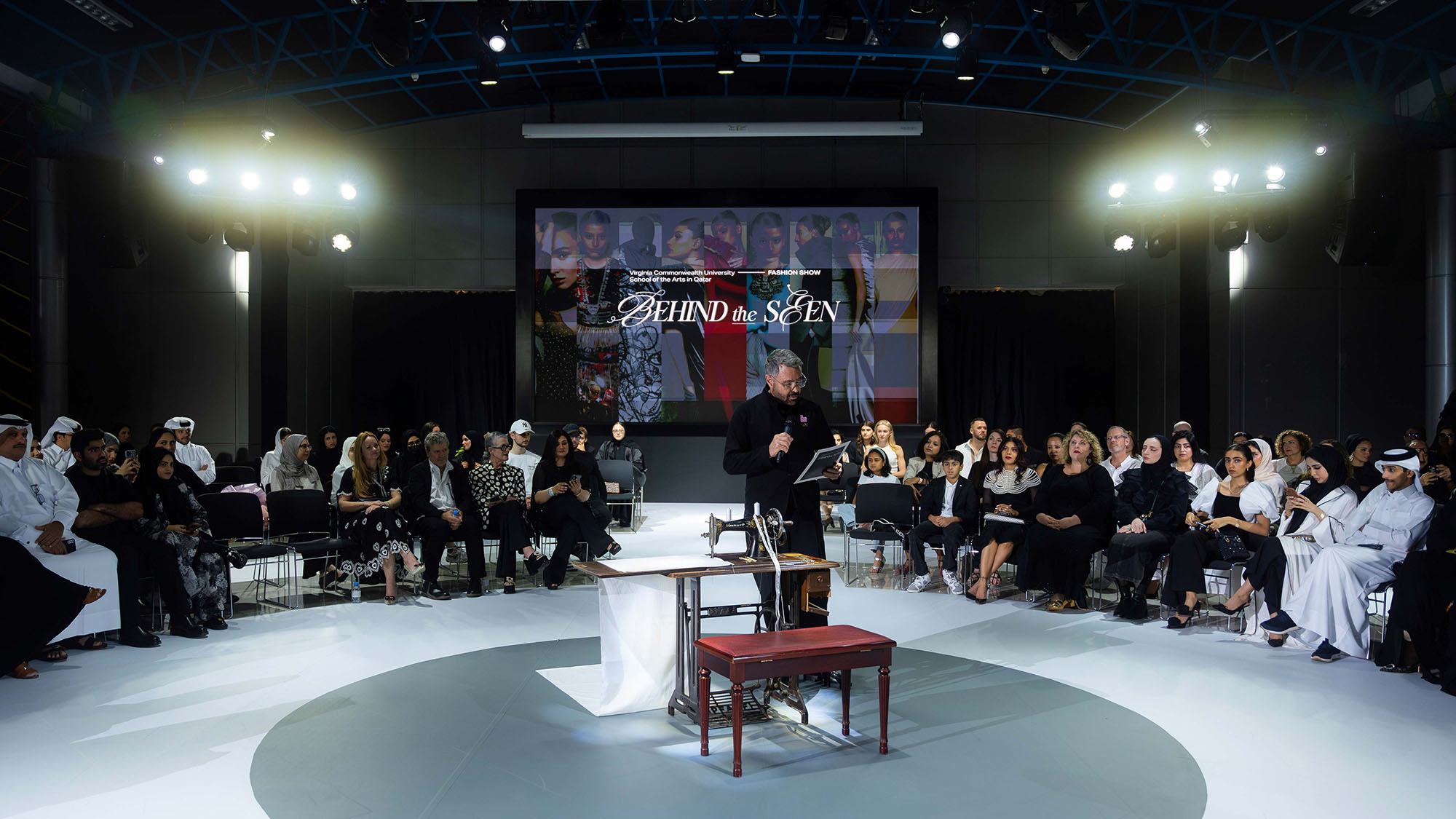Christopher Fink welcoming everyone to the fashion show . The runway is circular with two concentric rings made up of seats for the audience . In the center of the ring or runway is an old style sewing machine, and a stool , with a length of cloth hanging from it. In the background is a large video screen with the words behind the seen