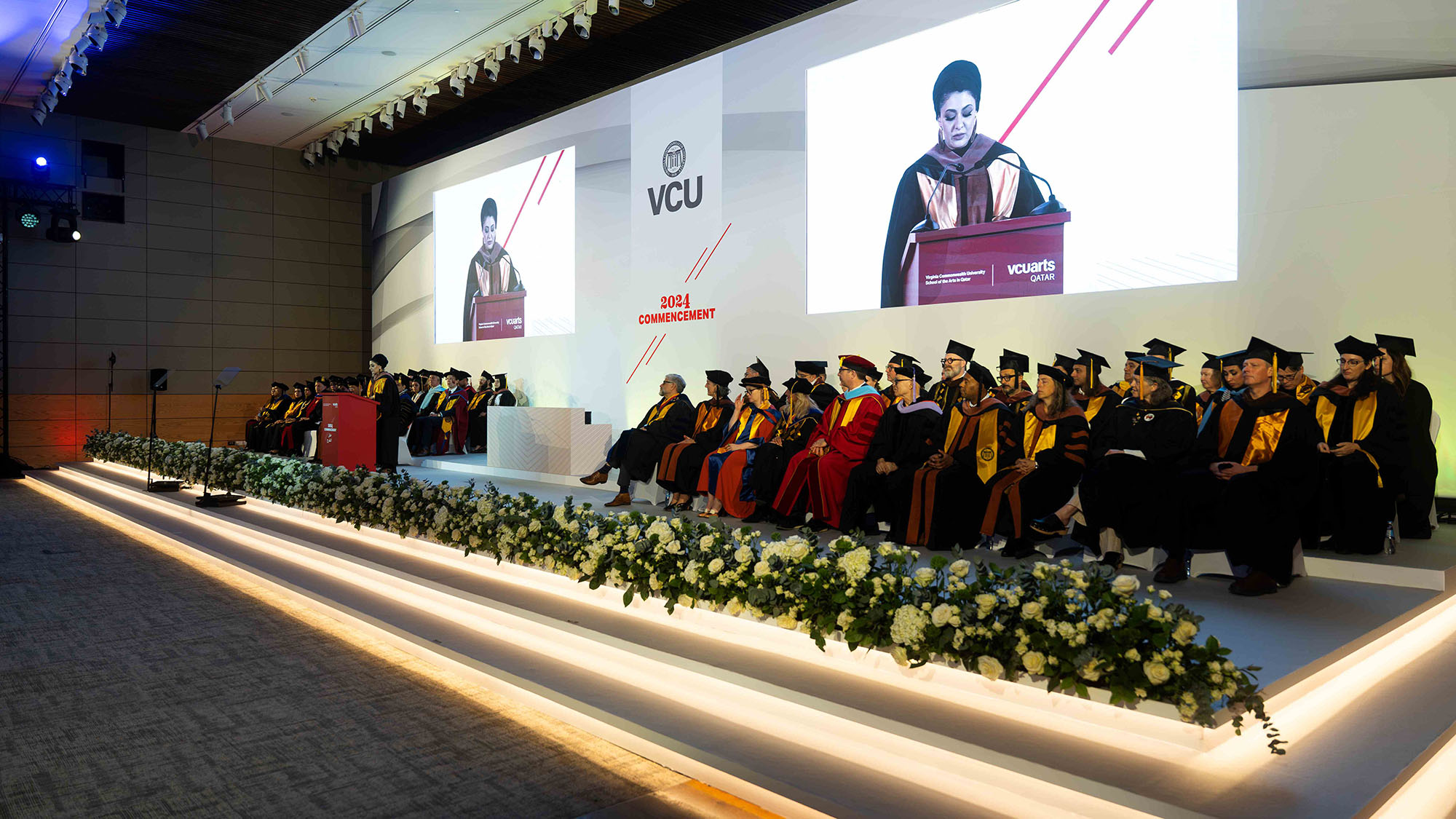 a Photo showing the platform party in the foreground with the keynote speaker in the background and on the screen above