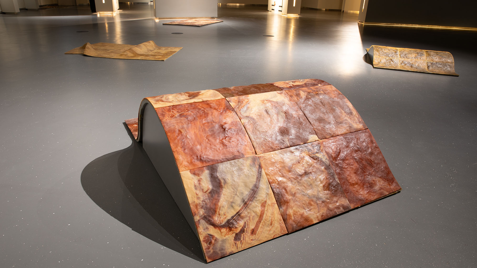 One of hana's work at the Wusum Exhibition . It is a wave shipped piece of wood constructed of squares . similar types of artwork are in the background . It is set against a gray floor