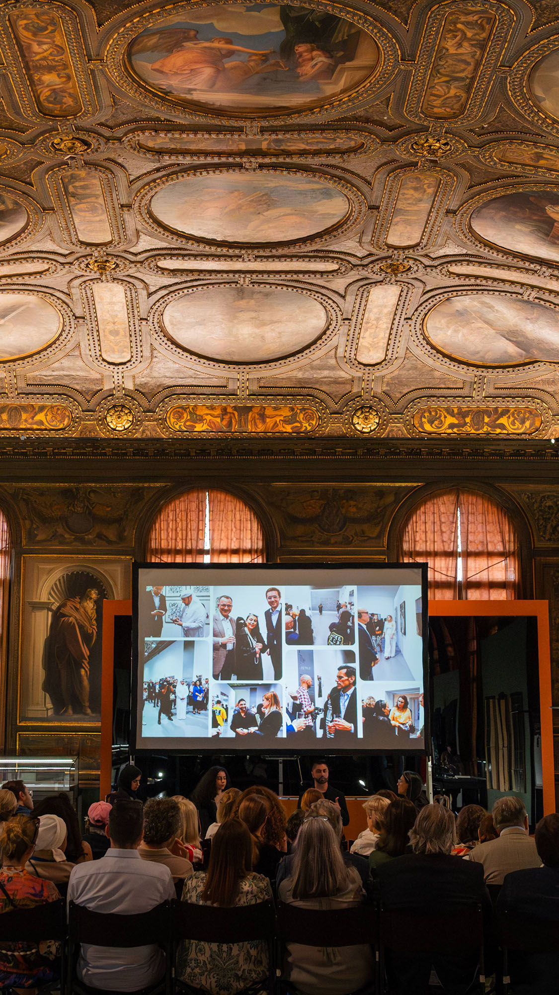 The interior of the the Biblioteca Marciana In Venice . It is an ornate auditorium with a richly decorated ceiling adorned with paintings and gold detailing. In the center, there’s a large screen displaying various images of individuals in professional and casual settings. On the stage below the screen, two presenters appear to be giving a lecture or presentation to an audience seated in rows of chairs. The setting suggests an event or conference blending historical architecture with modern technology.